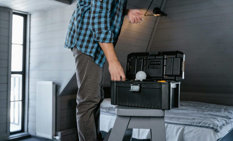 Person in plaid shirt fixing a light bulb using a toolbox in a rustic attic interior.