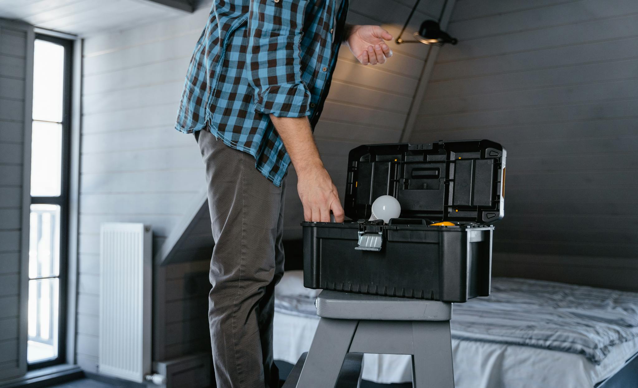 Person in plaid shirt fixing a light bulb using a toolbox in a rustic attic interior.